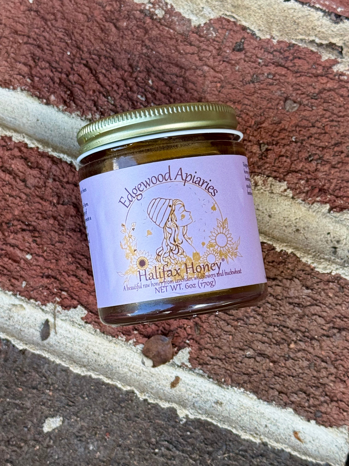 Halifax Honey 6 oz jar with a lavender label and gold lid, tilted against a red brick wall
