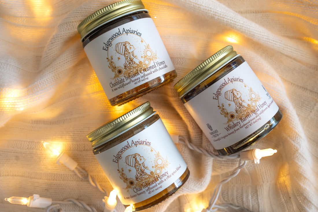 Three closed Edgewood Apiaries jars labeled Chocolate Infused Creamed Honey, Chai Infused Honey, and Whiskey Barrel Aged Honey rest on a cream knit blanket with warm string lights.