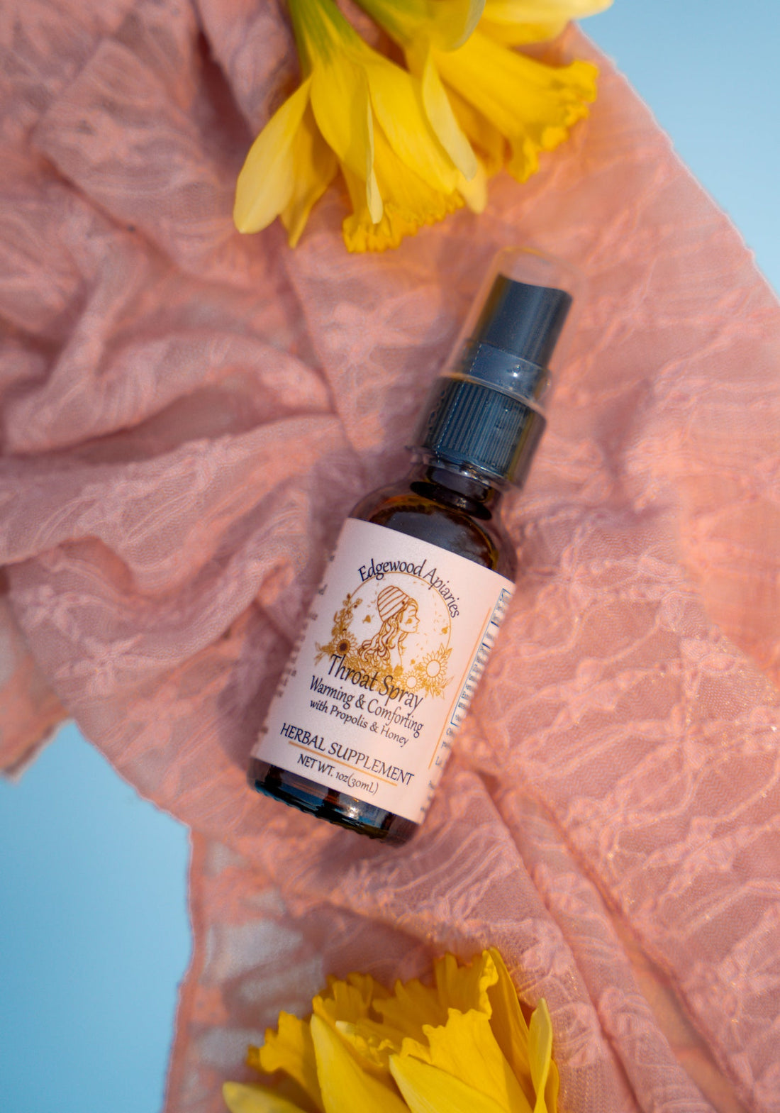 Edgewood Apiaries Warming & Comforting throat spray with propolis and honey. 1 oz amber bottle on pink lace with daffodils.
