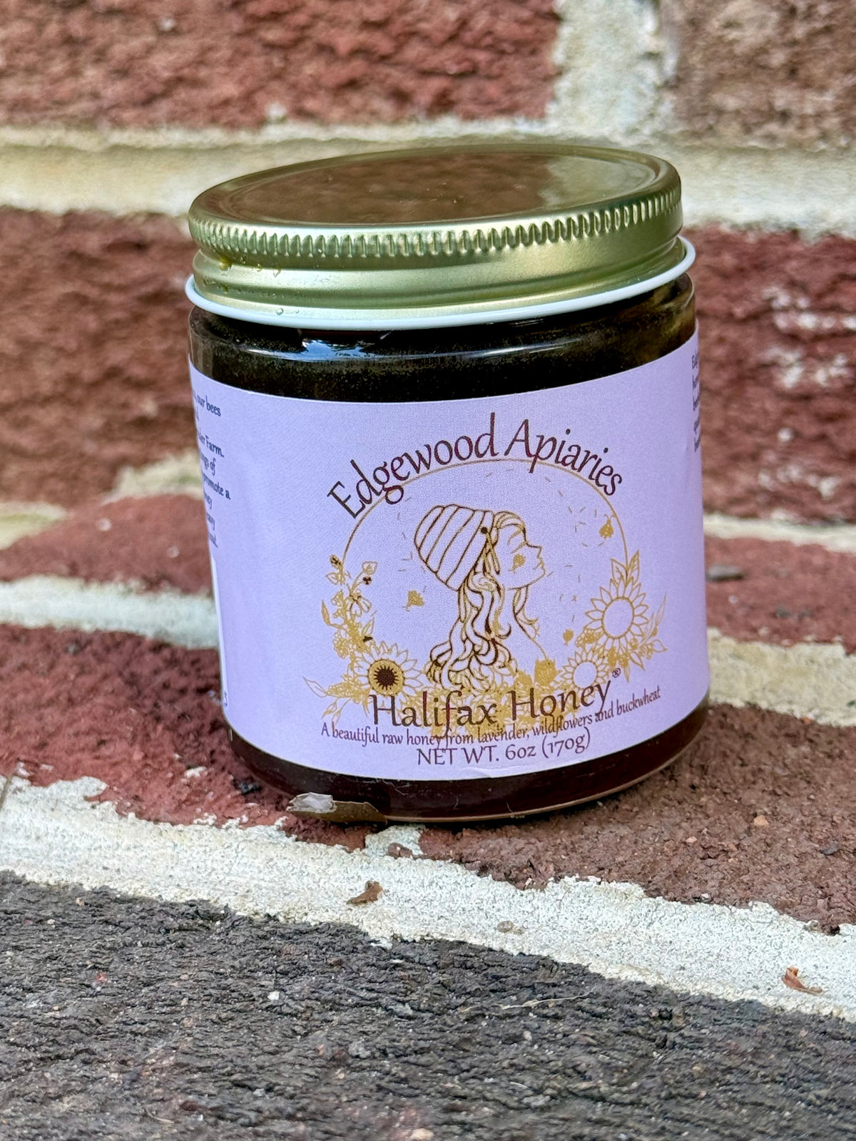 Halifax Honey 6 oz jar from Edgewood Apiaries with a gold lid and lavender label, standing on a brick ledge